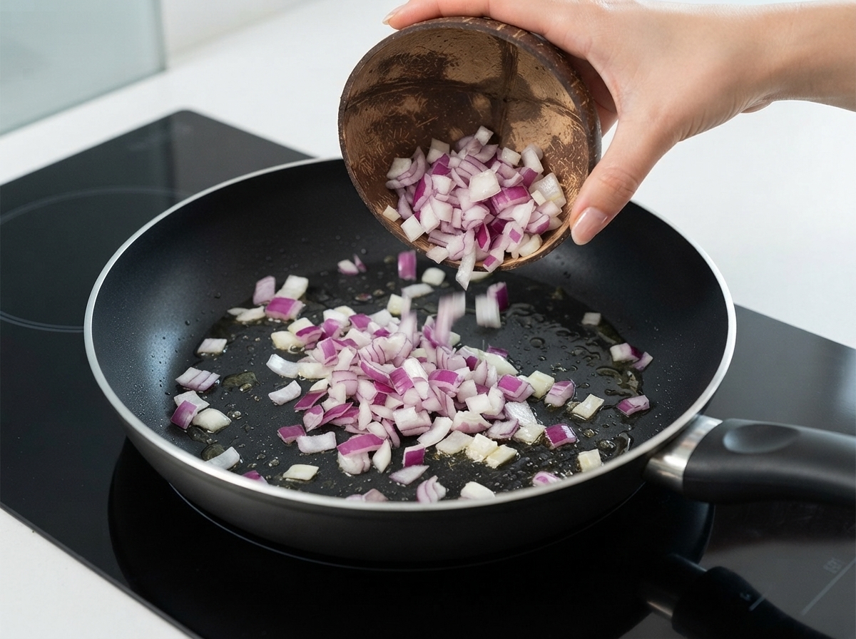 Cebollas rojas picadas siendo vertidas desde un bol de coco hacia una sartén con aceite.