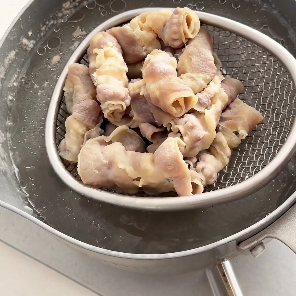 Un colador de malla de alambre levantando los rollitos de carne blanqueados de una olla de agua caliente.