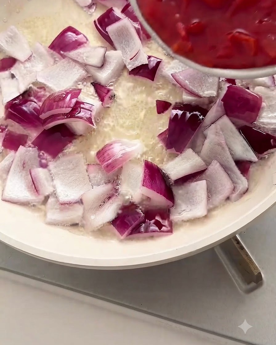 Tomates rojos picados siendo vertidos desde un tazón a una sartén blanca llena de cebolla morada picada y aceite.