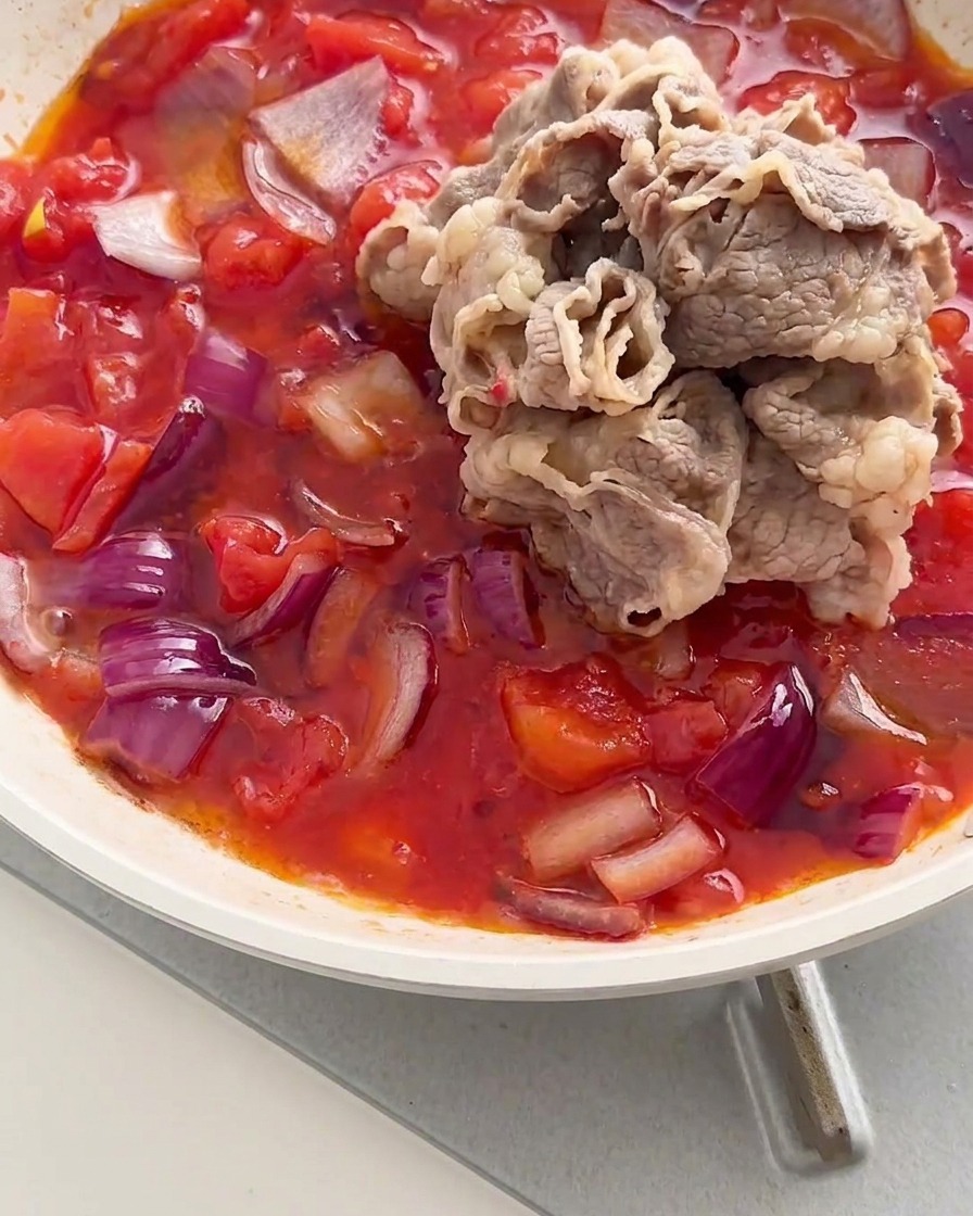 Una pila de rollitos de carne blanqueados añadidos a una sartén con una mezcla a fuego lento de tomates rojos y cebollas.