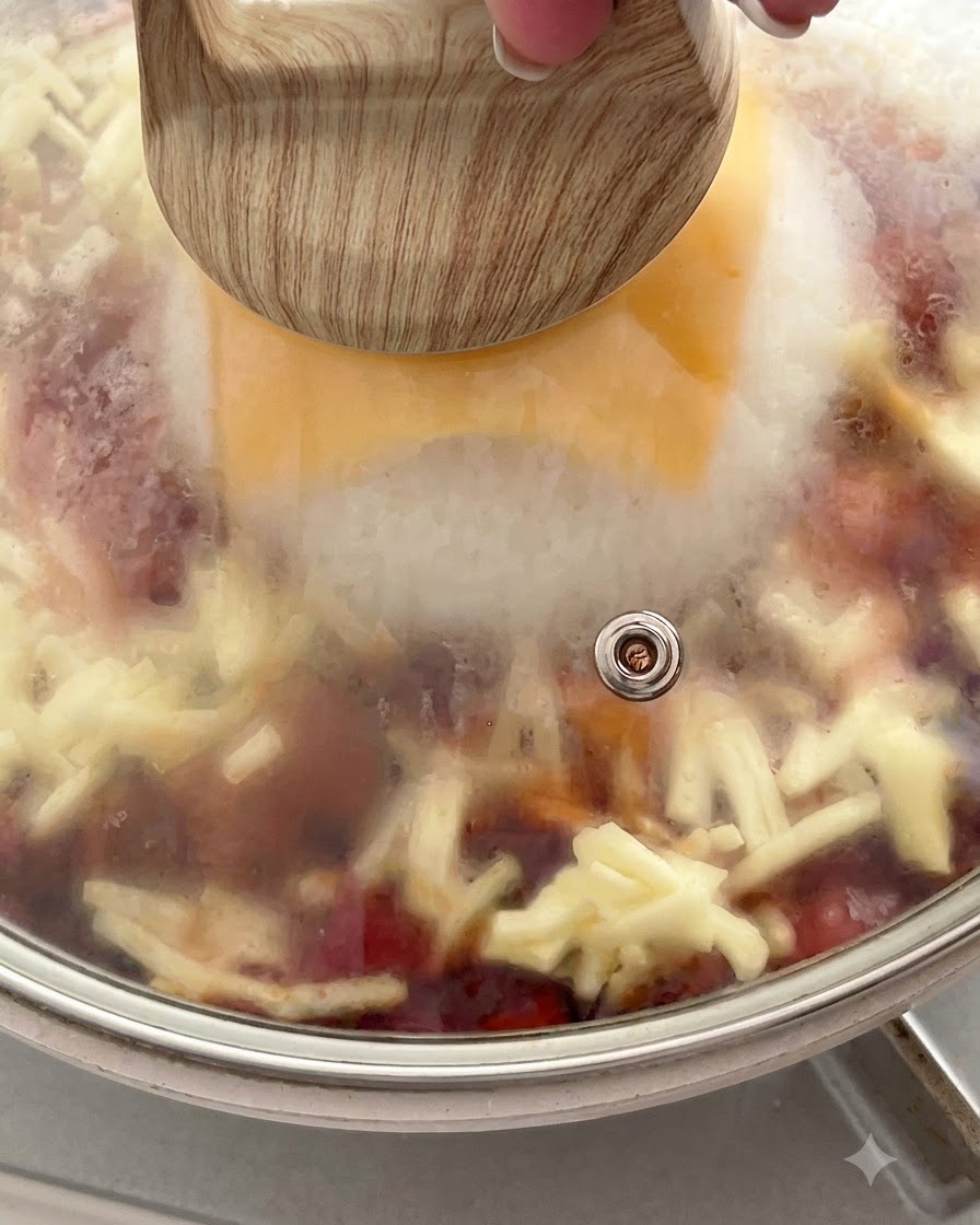 Una sartén cubierta con una tapa de vidrio que muestra vapor atrapado en su interior sobre el queso y el arroz derritiéndose.