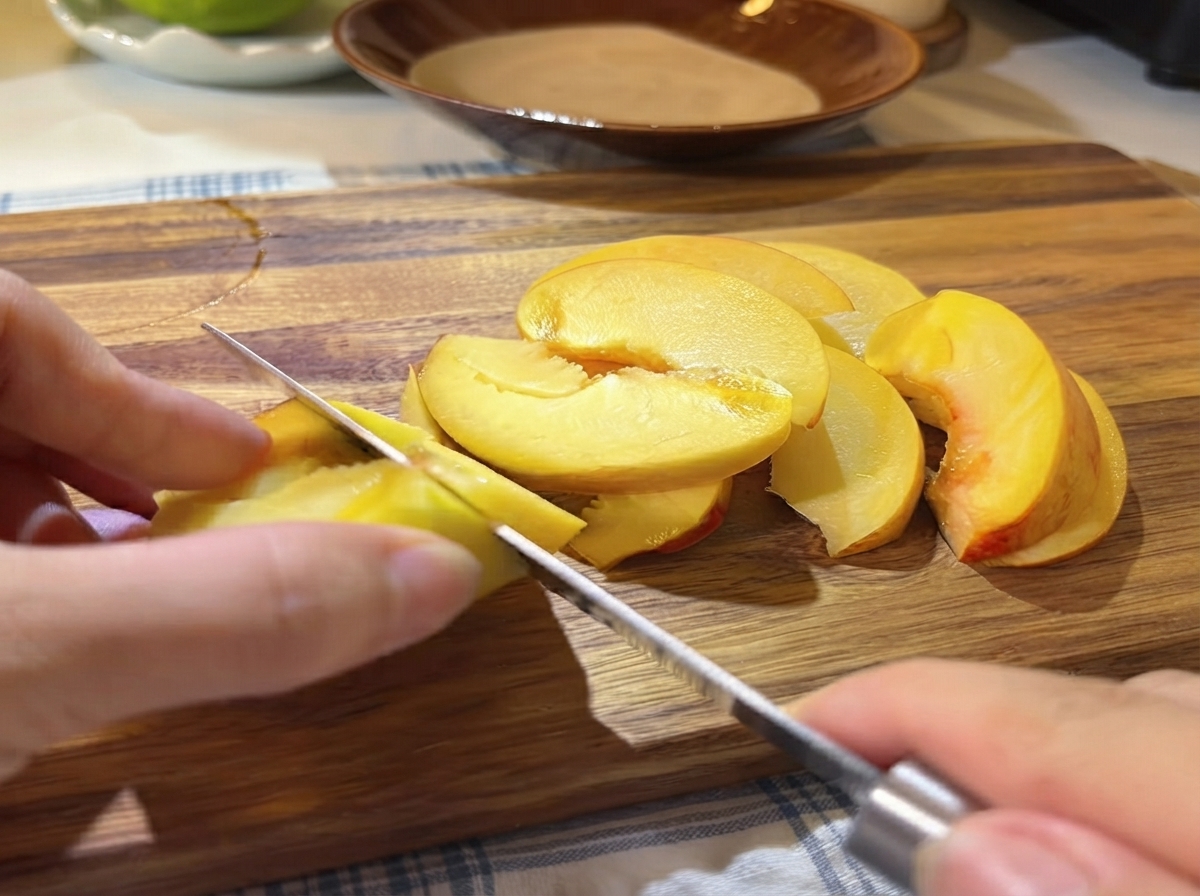 Una mano usando un cuchillo pequeño para cortar mitades de durazno amarillo fresco en rodajas finas sobre una tabla de cortar de madera.