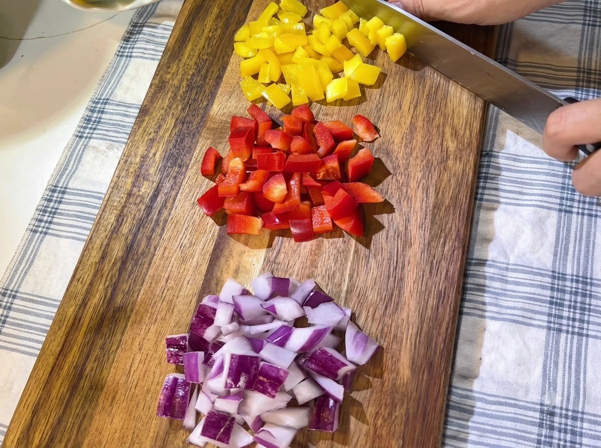 Montones de pimientos rojos y amarillos recién picados y cebolla morada en cubitos ordenados en una tabla de cortar de madera.