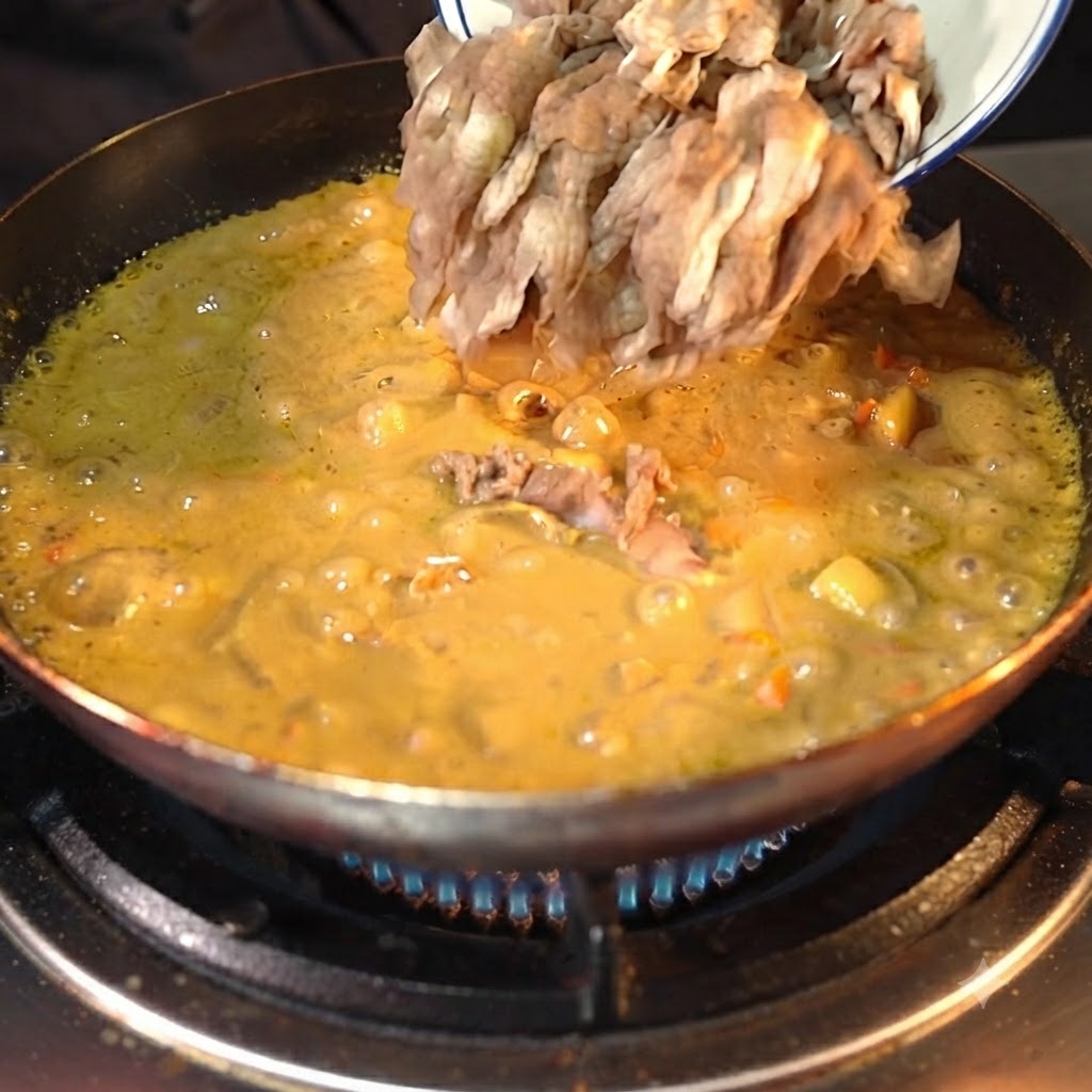 Trozos de ternera preprocesados siendo vertidos desde un cuenco blanco en una salsa de curry amarilla espesa y burbujeante en una sartén oscura.