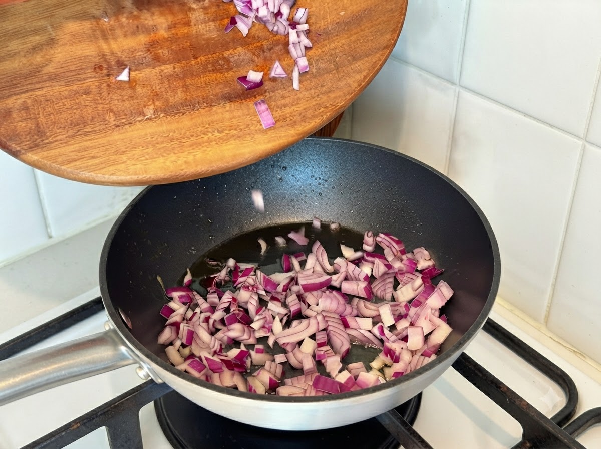 Cebollas rojas picadas siendo vertidas desde una tabla de madera a una sartén caliente con aceite.