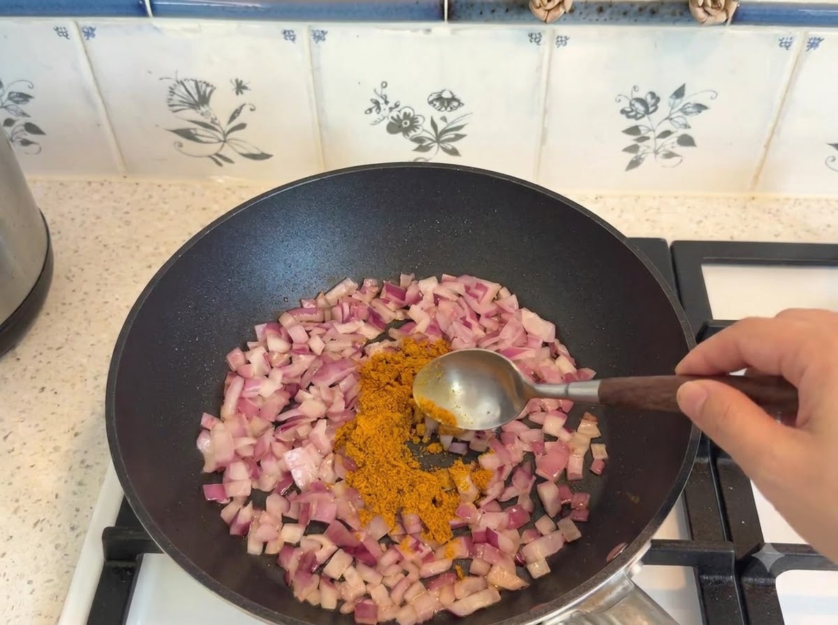 Polvo de curry siendo medido con una cuchara y añadido a cebollas salteadas en una sartén.