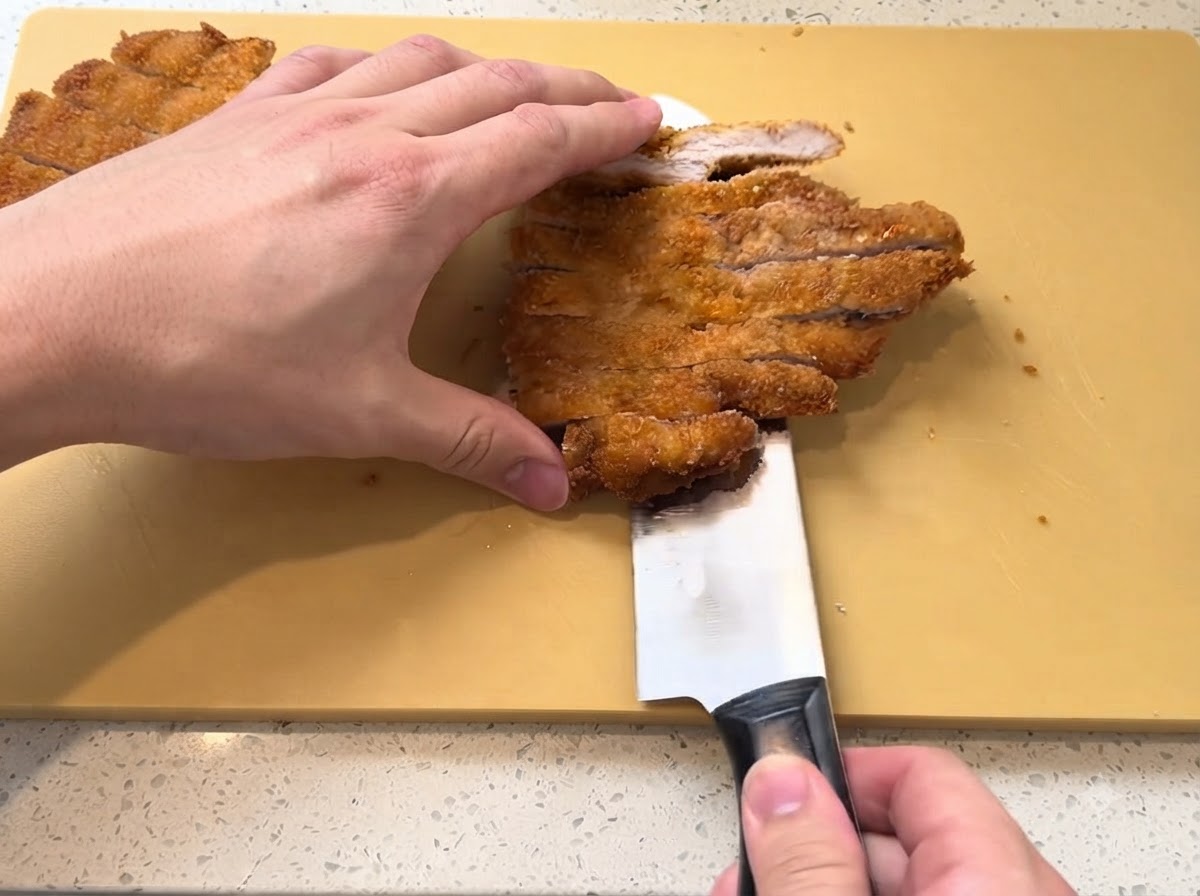 Un filete de cerdo frito de color marrón dorado siendo cortado en tiras con un cuchillo afilado en una tabla de cortar amarilla.