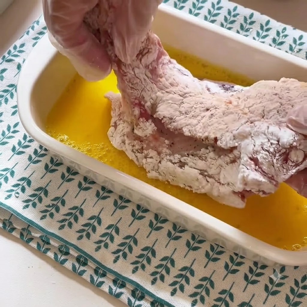 Una mano con guante levantando una chuleta de cerdo cubierta de almidón de un recipiente con líquido de huevo batido amarillo.