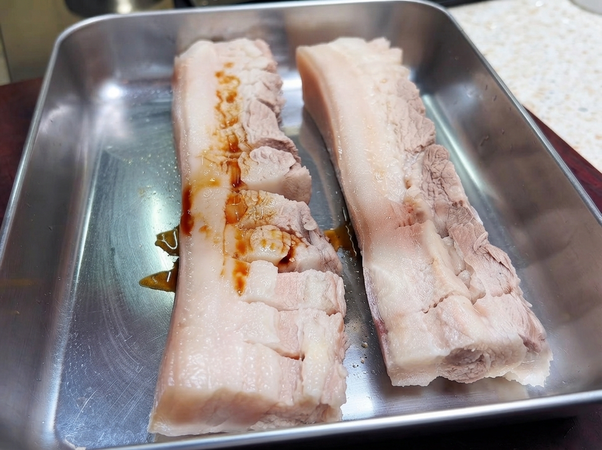 Dos trozos de panceta con cortes en una bandeja de metal con salsa de soja oscura vertida sobre la carne.