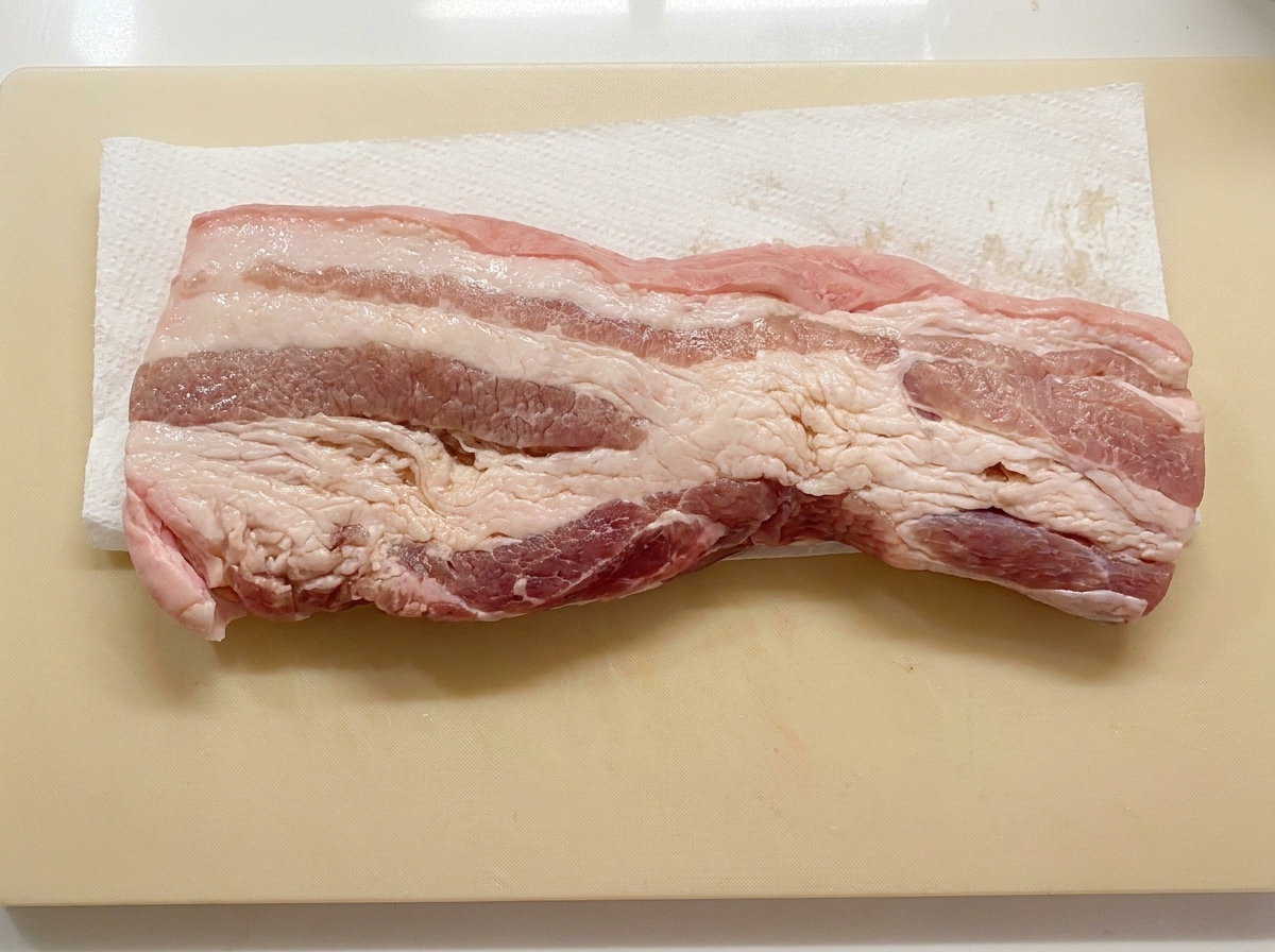 Una tira de panceta de cerdo colocada plana sobre una toalla de papel en una tabla de cortar mientras se seca.