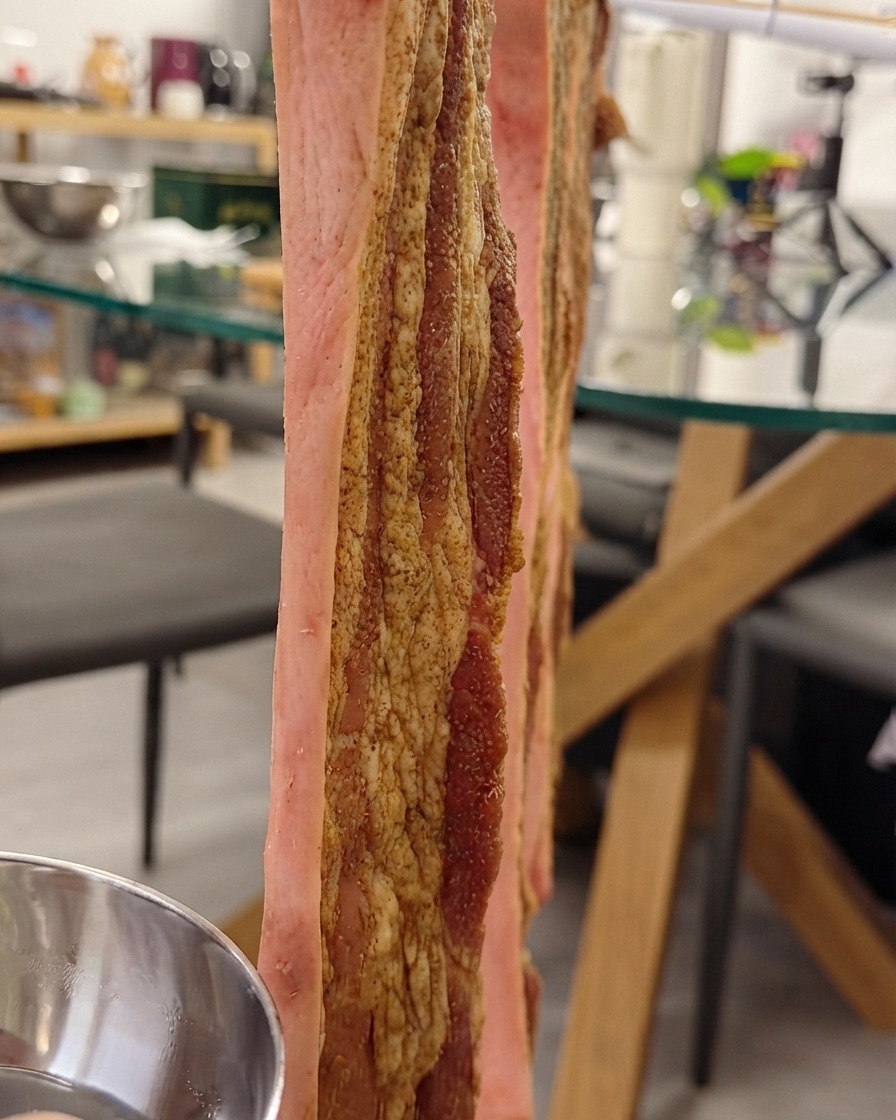 Panceta de cerdo colgando verticalmente en una rejilla mientras se le cepilla agua para la piel crujiente en la piel.