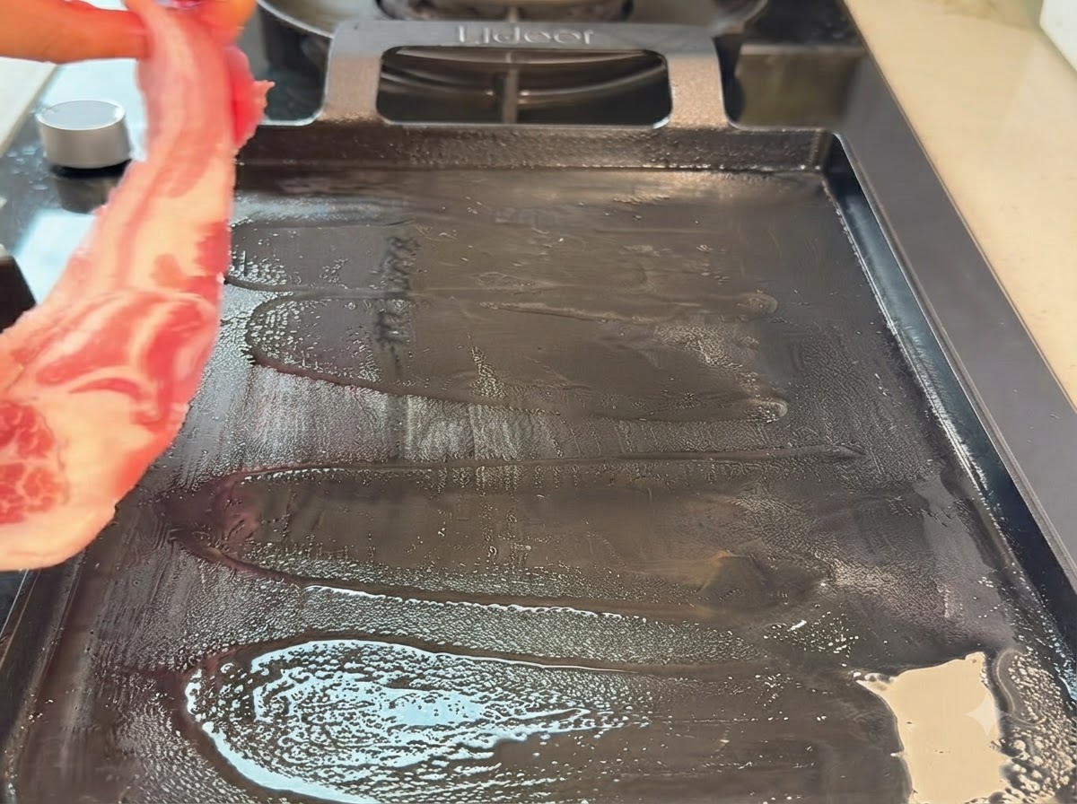 Una tira de panceta de cerdo cruda colocándose en una sartén grill teppanyaki caliente.