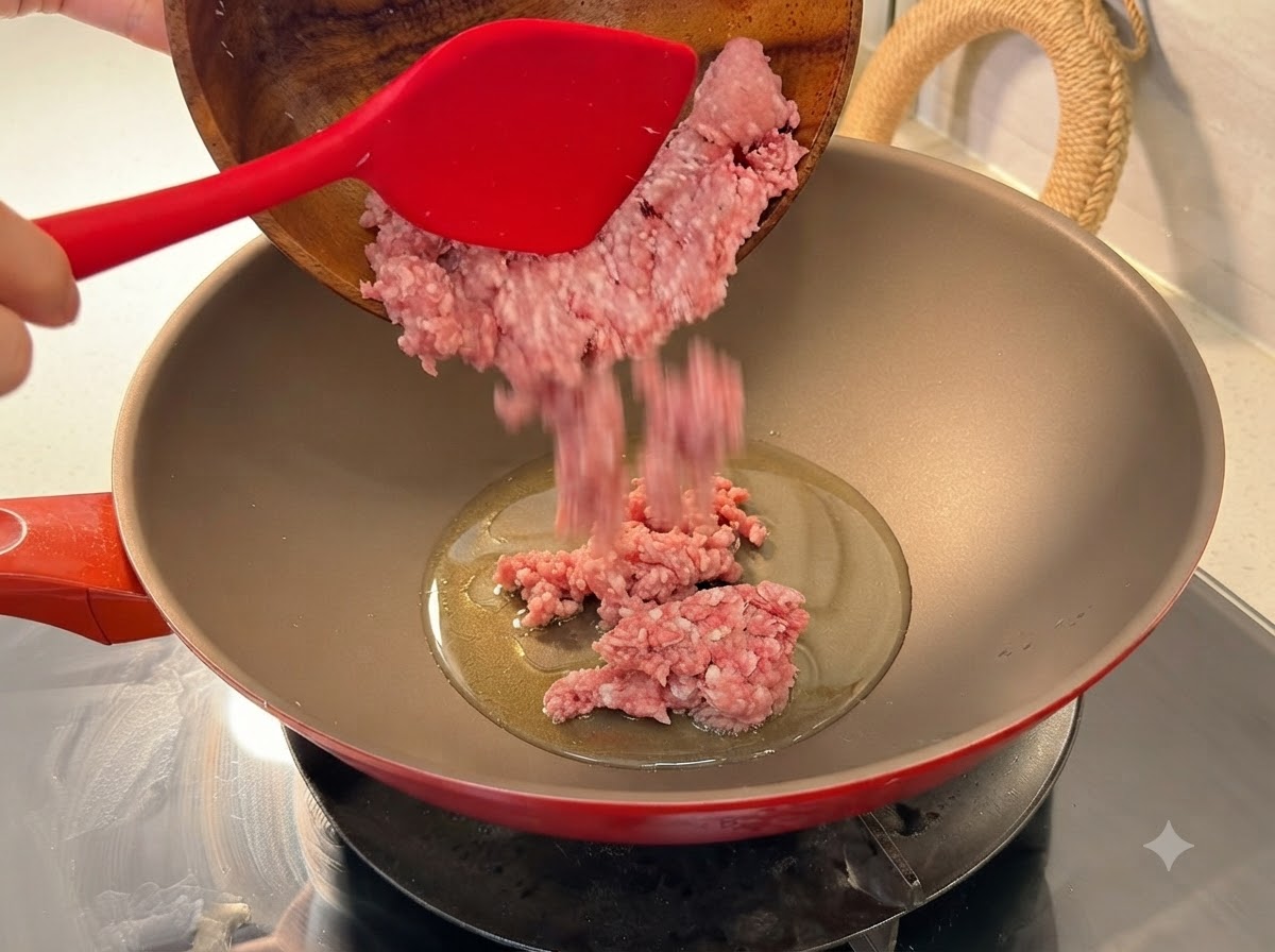 Carne de cerdo picada fresca vertida desde un bol de madera en un wok rojo caliente con aceite.