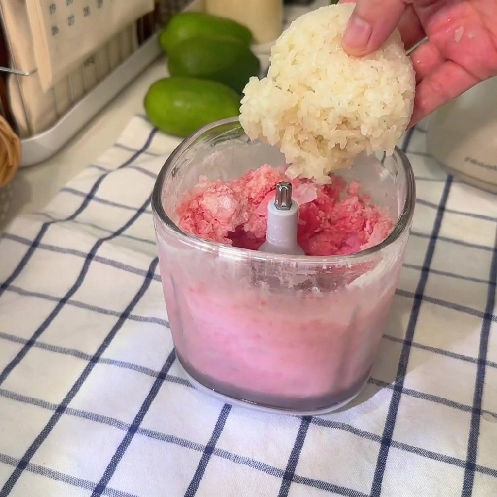 Una bola de arroz glutinoso cayendo en un vaso de licuadora transparente lleno de mezcla rosa de granizado de sandía.