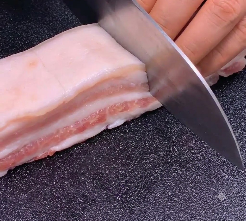 Un cuchillo afilado haciendo cortes precisos y uniformes en una tira de panceta de cerdo sobre una tabla de cortar oscura.