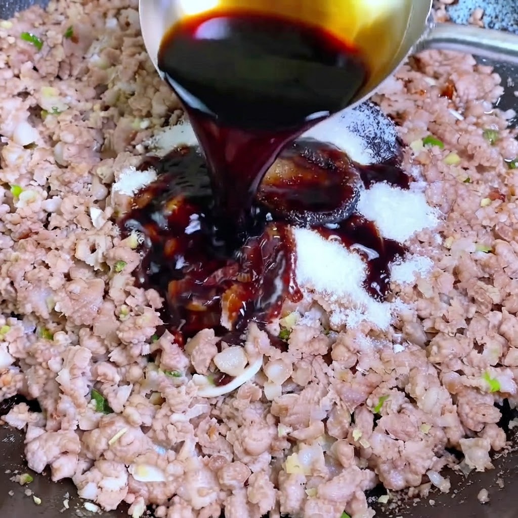 Salsa de soja oscura siendo vertida desde un cucharón plateado a un wok que contiene carne de cerdo picada y condimentos blancos.