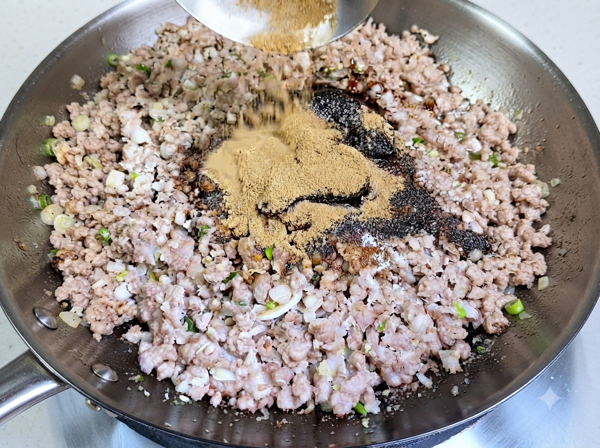 Un montón generoso de polvo de especias marrón claro siendo vertido desde un cucharón al wok sobre la carne de cerdo picada sazonada.