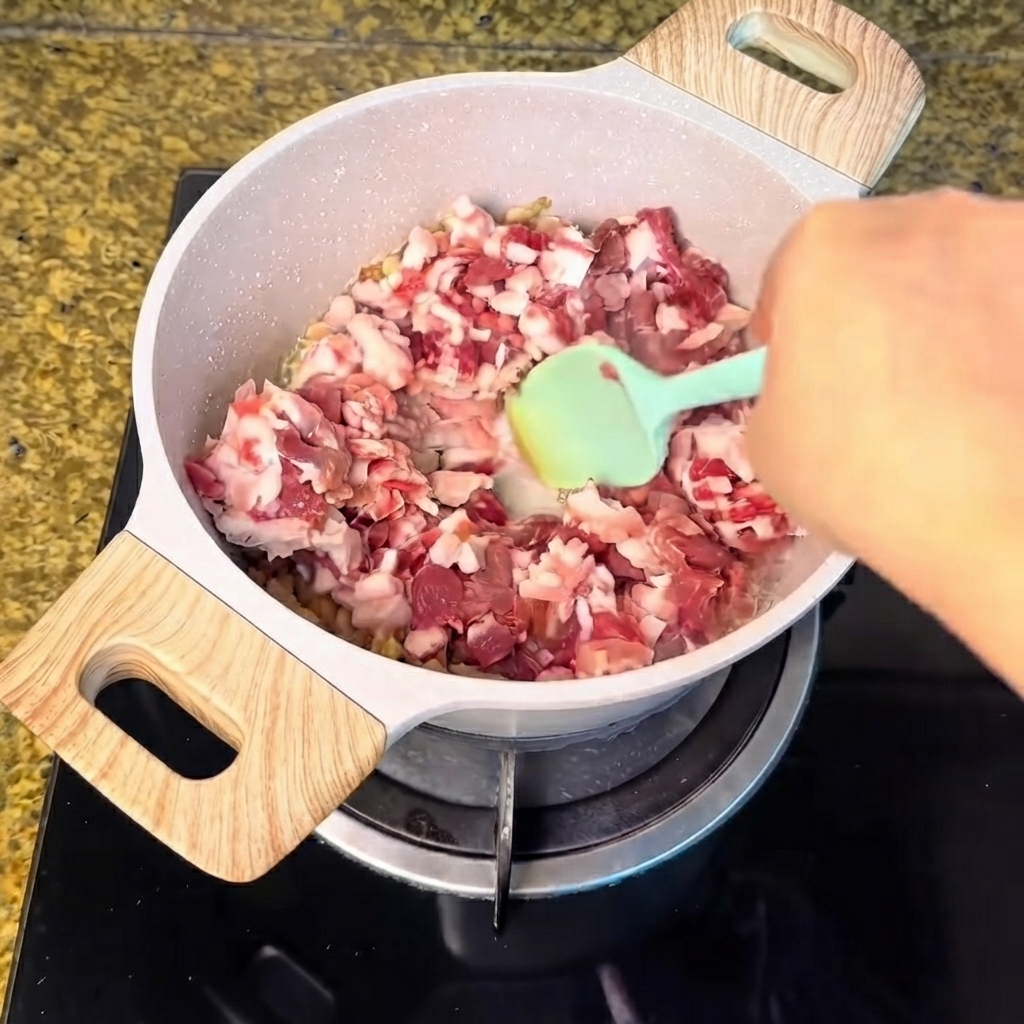 Carne de cerdo en dados vertida en la olla con grasa fundida y mezclada con una espátula verde.