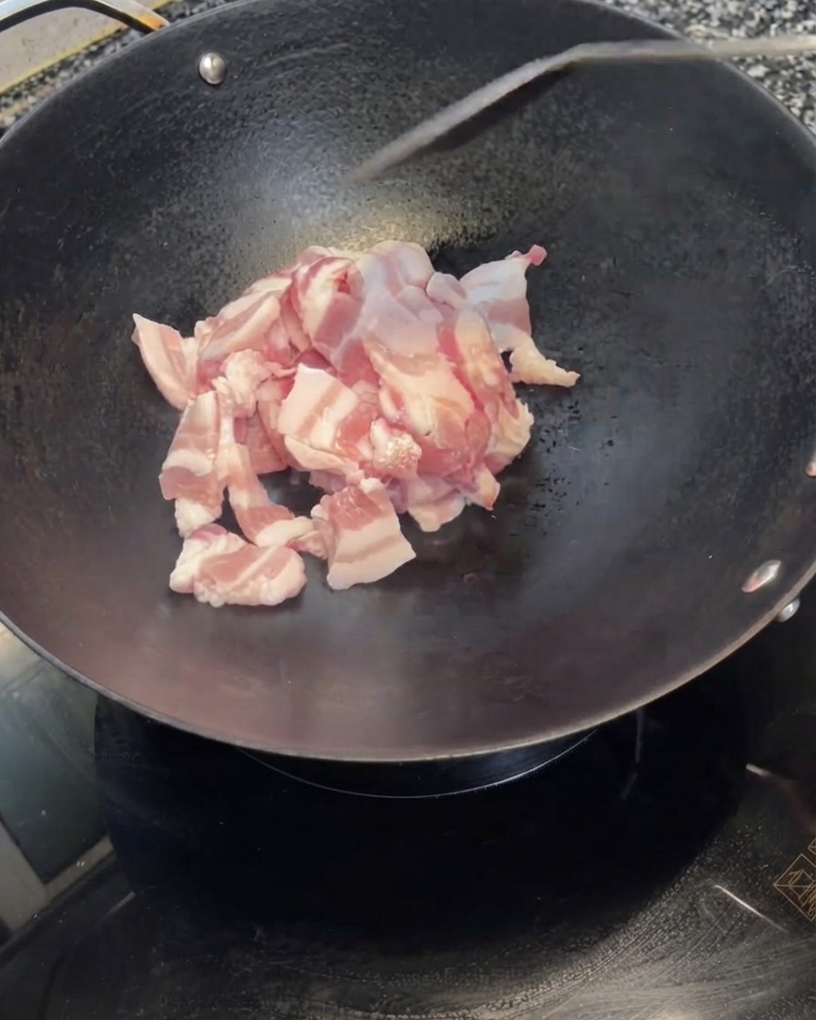 Rodajas de panza de cerdo siendo vertidas desde un tazón blanco en un wok negro caliente.