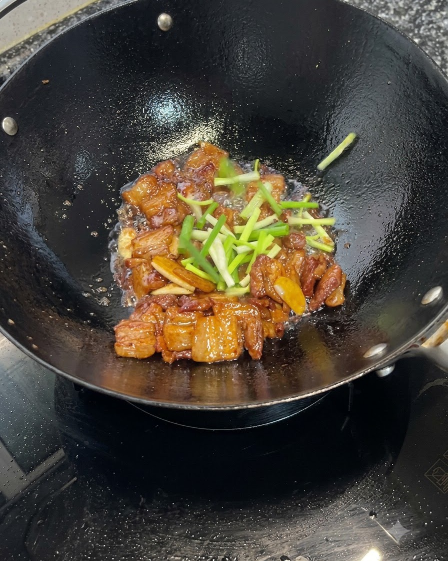 Secciones de cebollino fresco esparcidas sobre trozos de panza de cerdo caramelizada cocinándose en un wok.