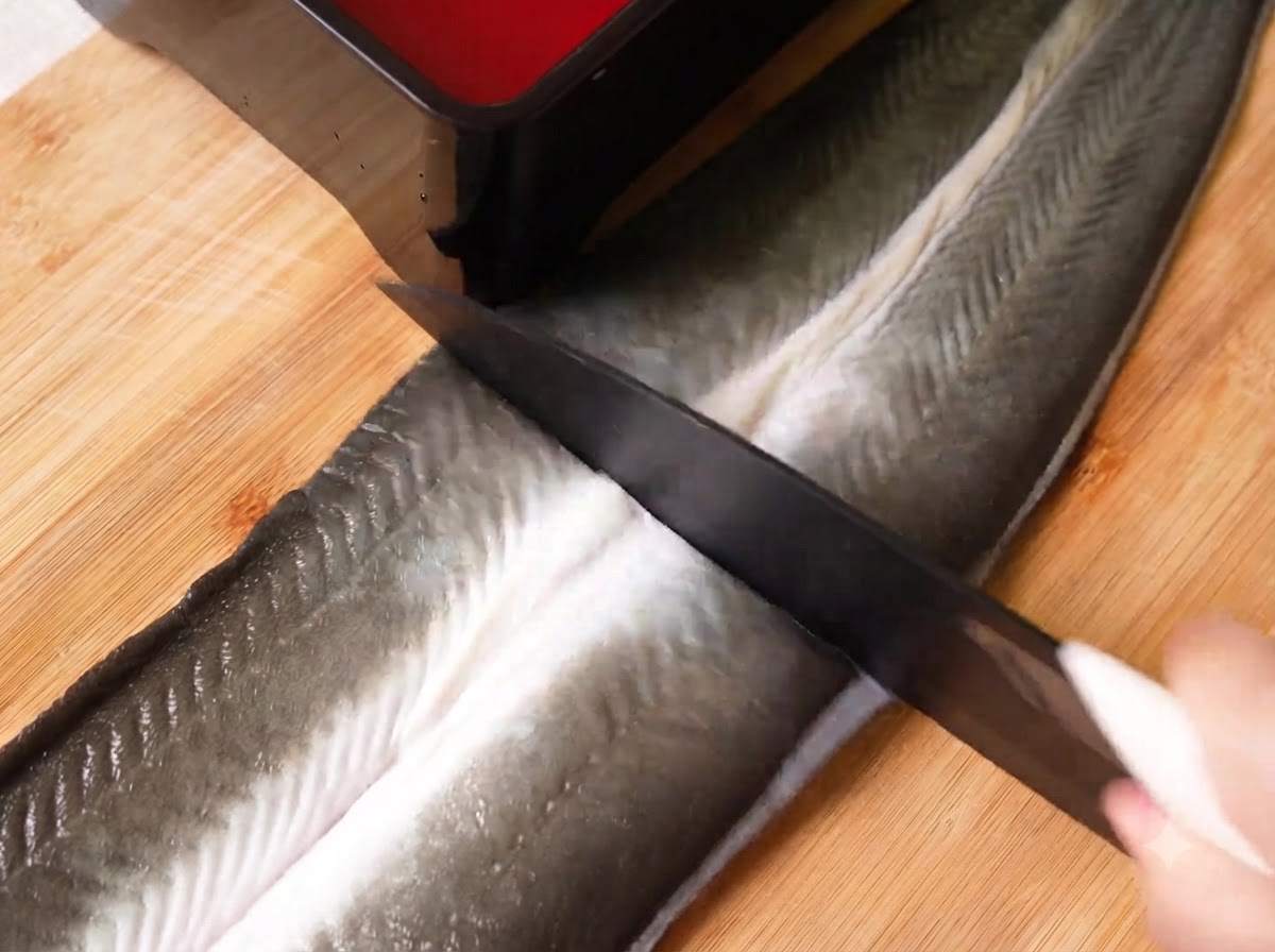 Un cuchillo afilado cortando una gran anguila cruda en mariposa en tres secciones sobre una tabla de cortar de madera.