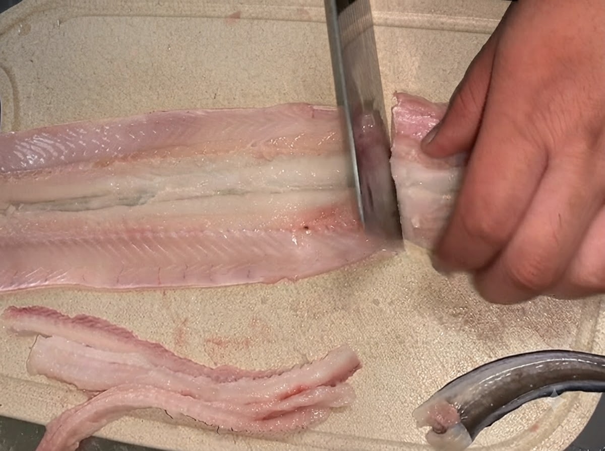 Un cuchillo cortando un filete de anguila cruda desespinada en dos secciones separadas sobre una tabla de cortar.