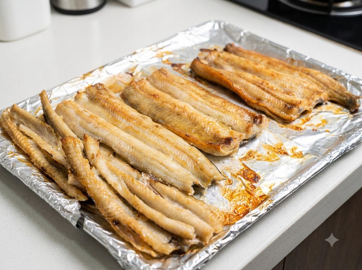 Una vista de cerca de los filetes de anguila parcialmente horneados siendo pincelados con aceite de cocina sobre una bandeja de horno.