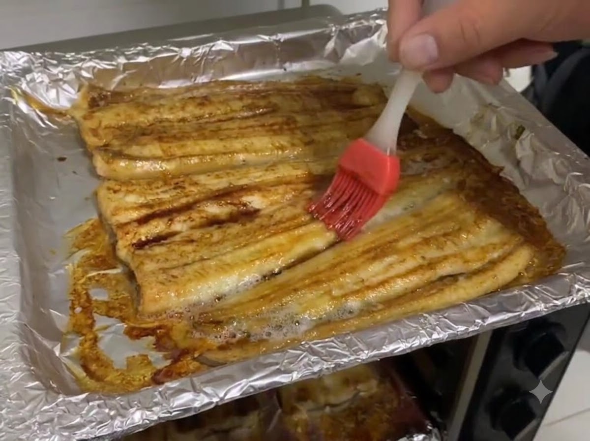Salsa de soya oscura siendo aplicada con brocha sobre la superficie de los filetes de anguila horneados en una bandeja cubierta con papel aluminio.