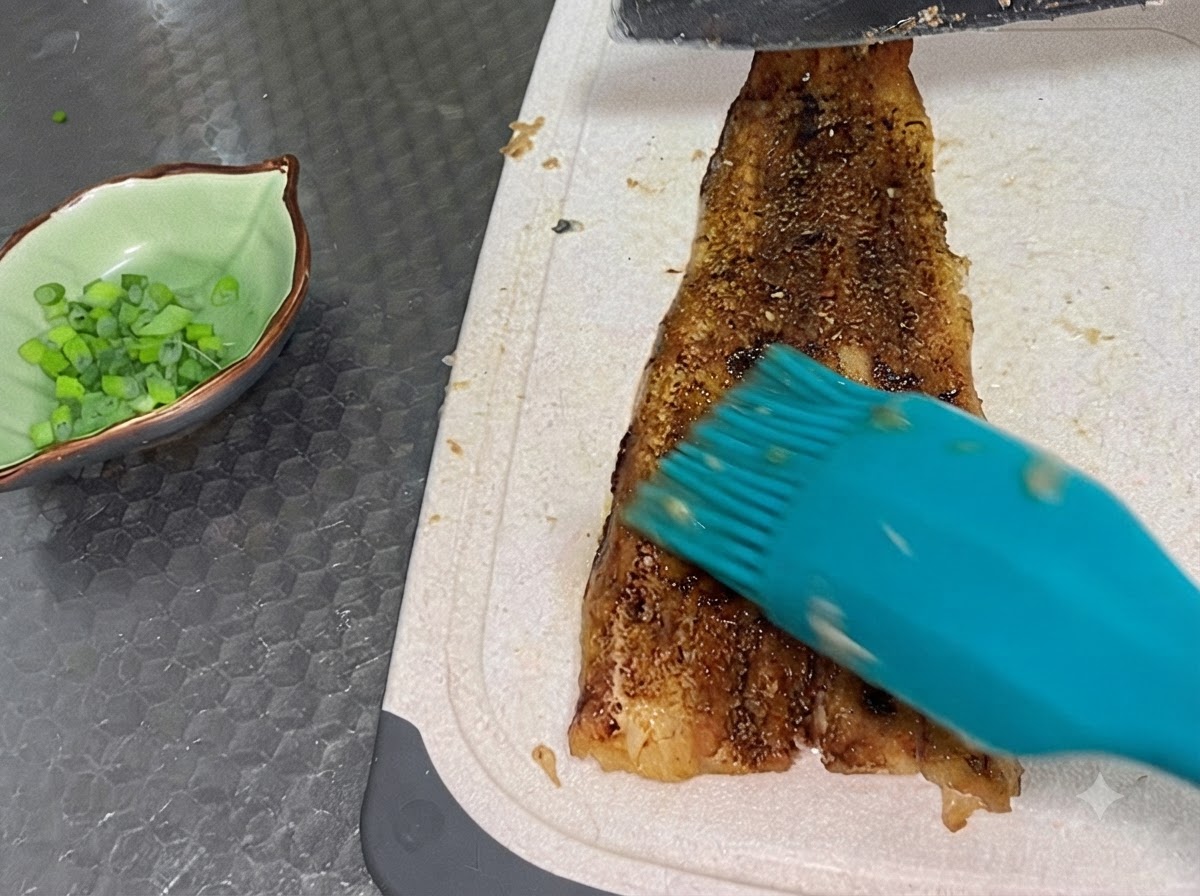 Un filete de anguila completamente horneado siendo cortado en una tabla de cortar blanca junto a un pequeño tazón verde lleno de cebollines picados.