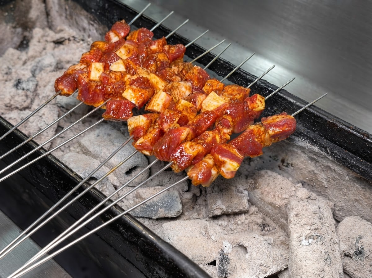 Múltiples brochetas de metal con carne de cerdo sazonada dispuestas ordenadamente sobre una parrilla de carbón encendida.