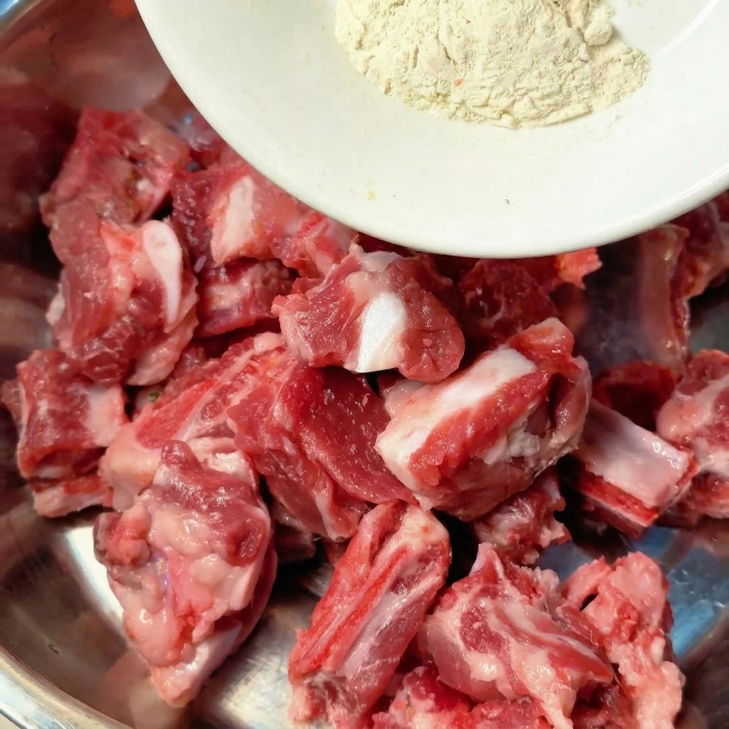 Especias y condimentos líquidos siendo vertidos directamente sobre costillas de cerdo crudas en un tazón de metal para mezclar.