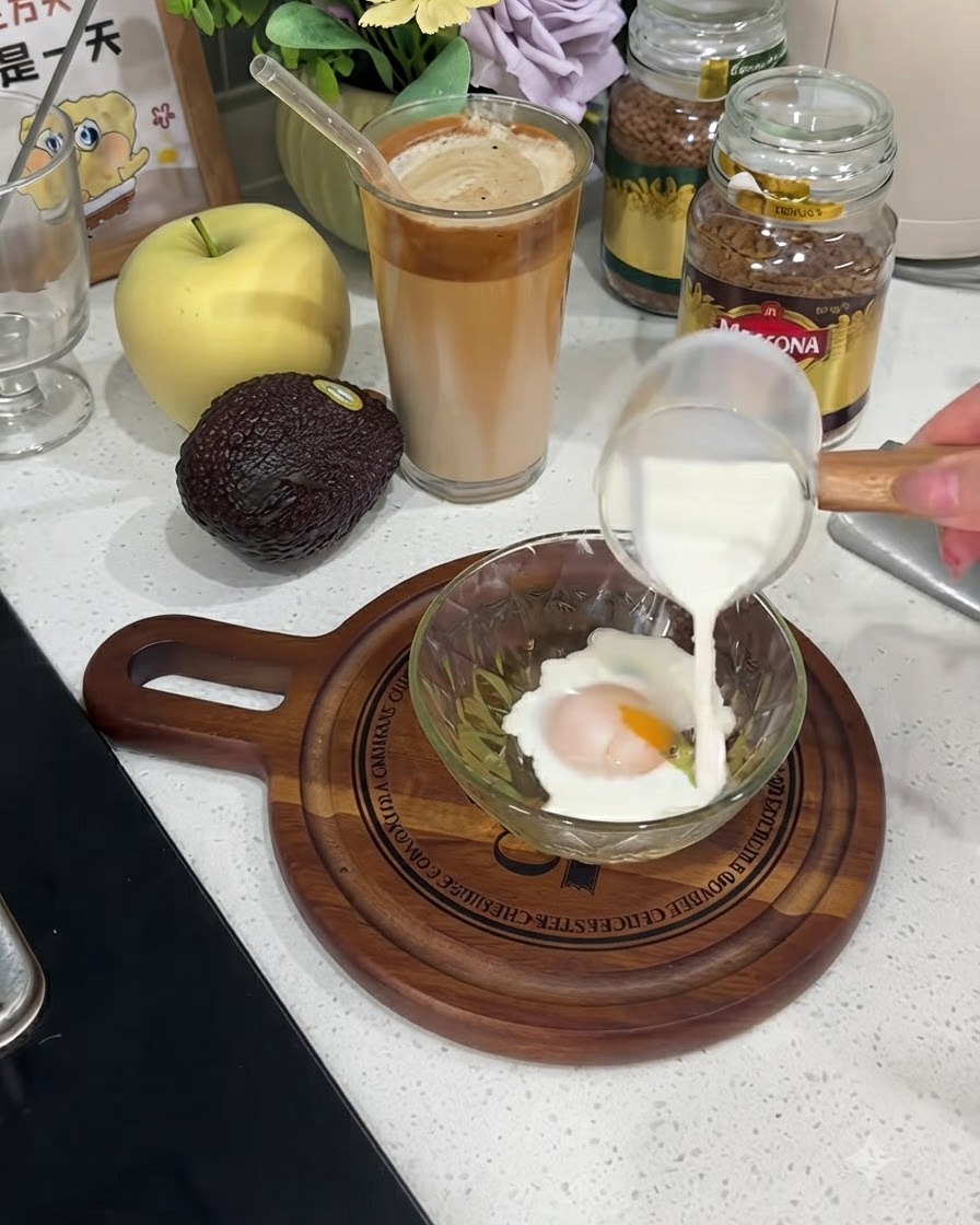 Leche siendo vertida desde una pequeña jarra de vidrio hacia un tazón que contiene un huevo roto.