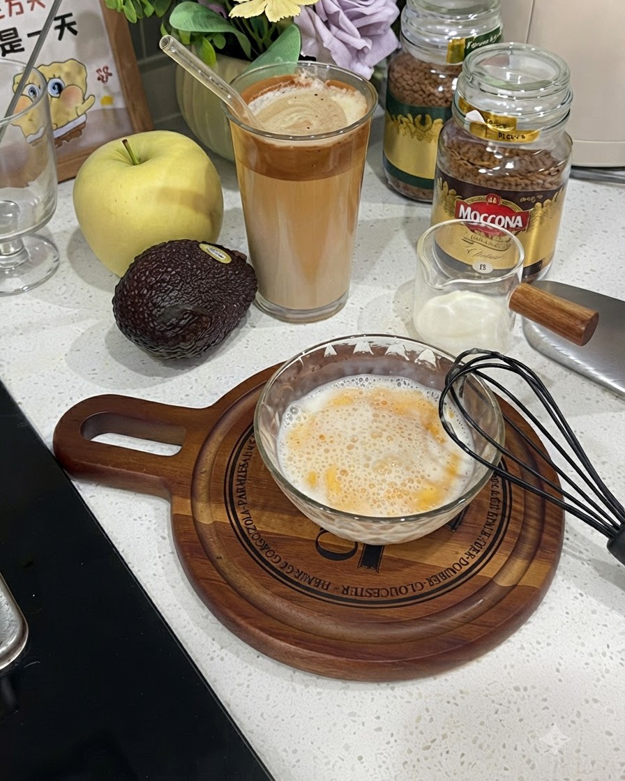 Un tazón de vidrio lleno de una mezcla batida y espumosa de huevo y leche al lado de un batidor negro.