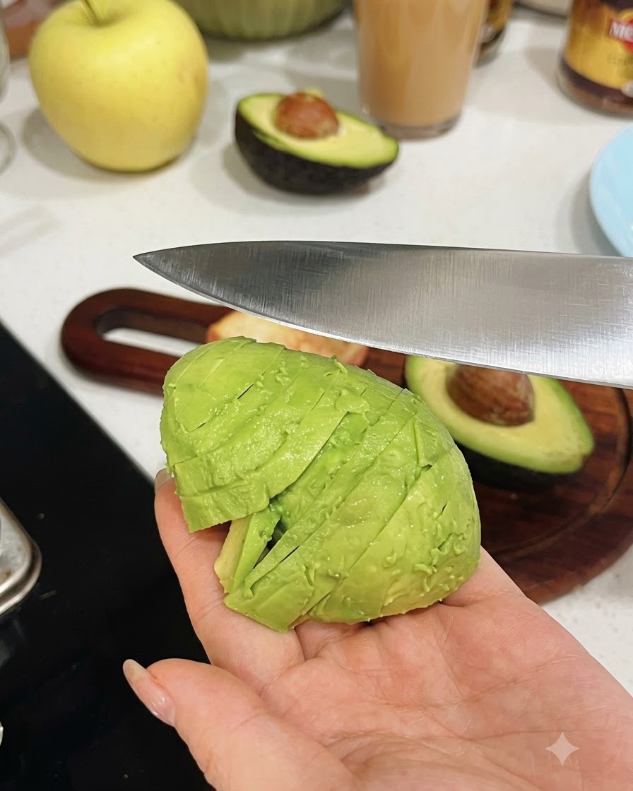 Una mano sosteniendo una mitad de aguacate pelado mientras un cuchillo de cocina lo corta en piezas uniformes.