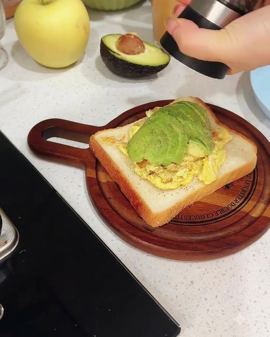 Un molinillo de pimienta moliendo pimienta negra fresca directamente sobre una rebanada de tostada cubierta con huevos revueltos y aguacate en rodajas.