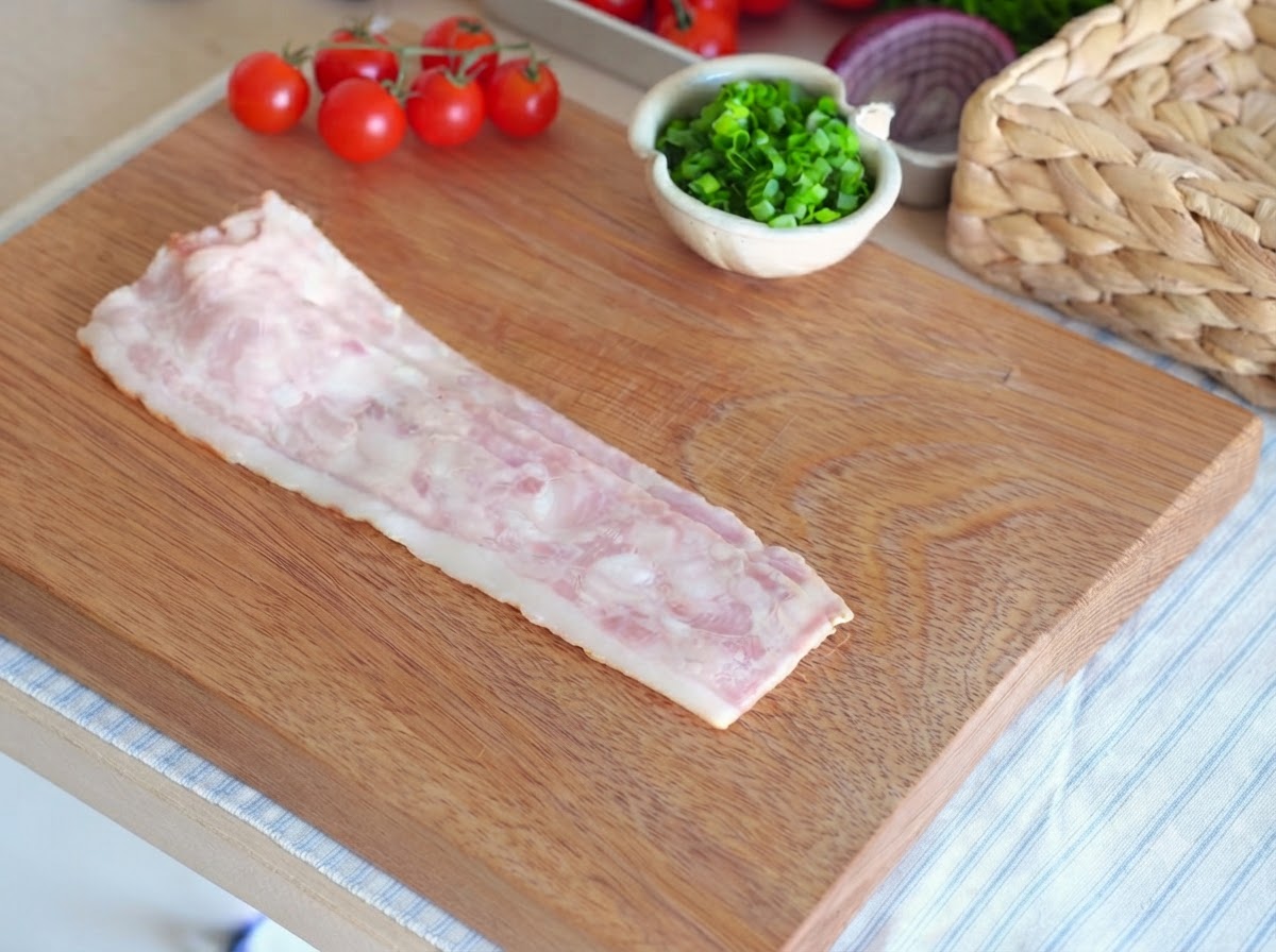 Una sola tira de tocino crudo extendida sobre una tabla de cortar de madera lista para ser cortada.