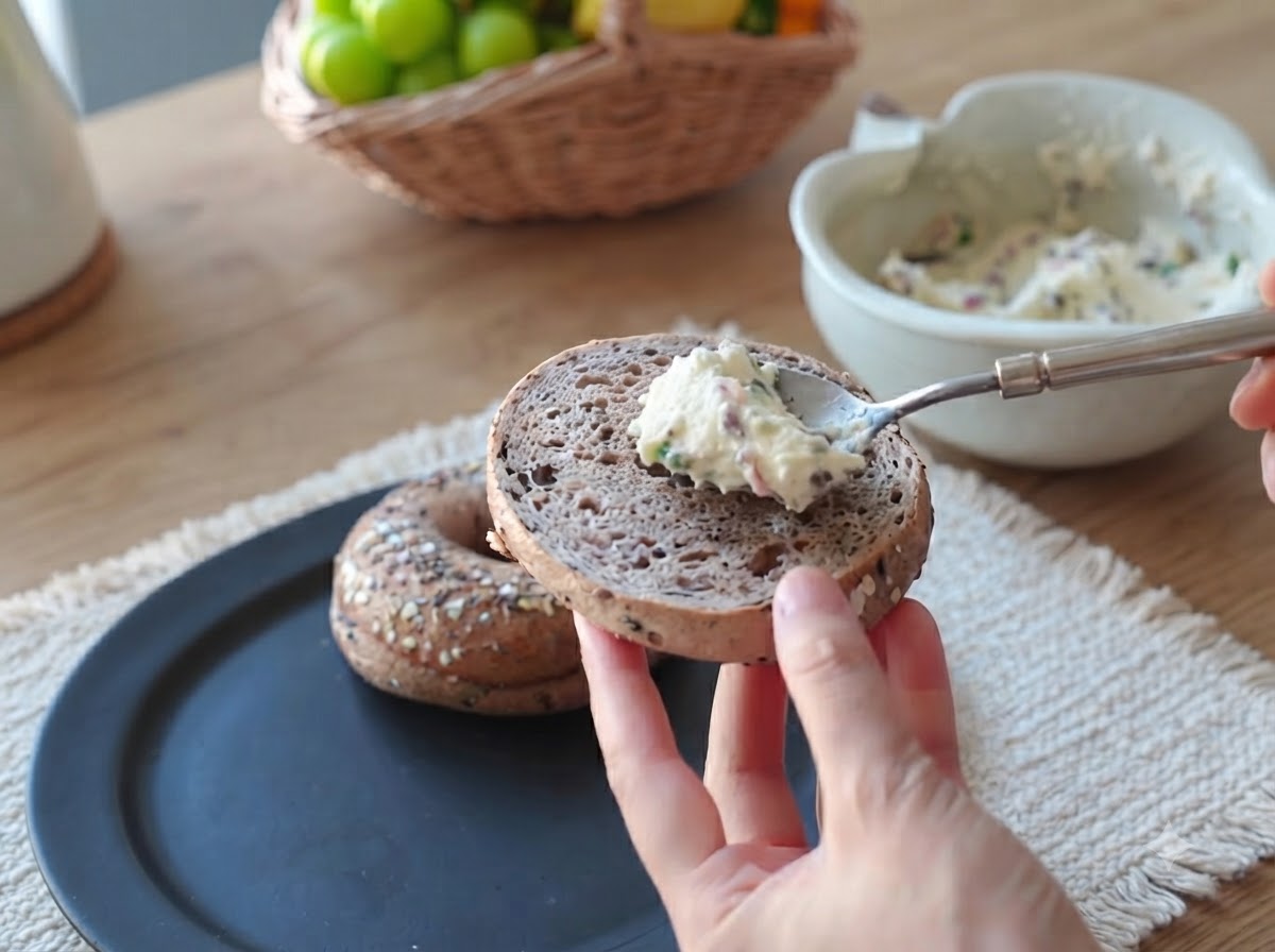 Una cucharada de crema untable de tocino y cebollino siendo alisada sobre la superficie de la mitad de un bagel.