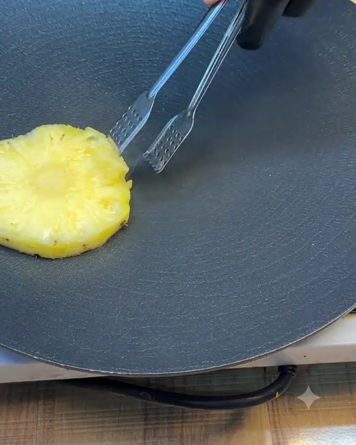 Una sola rodaja de piña siendo girada en una plancha oscura con pinzas de metal.
