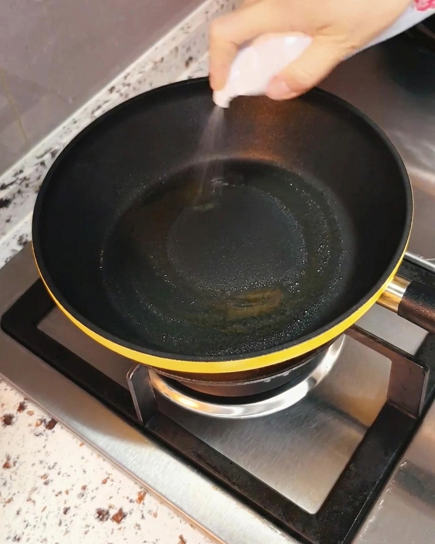 Una mano rociando aceite de cocina de manera uniforme en una sartén negra colocada sobre la hornalla de una estufa.