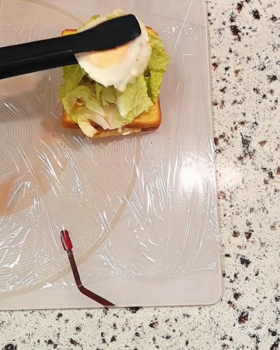 Un par de pinzas negras sosteniendo un huevo cocido sobre una capa de sándwich con lechuga.