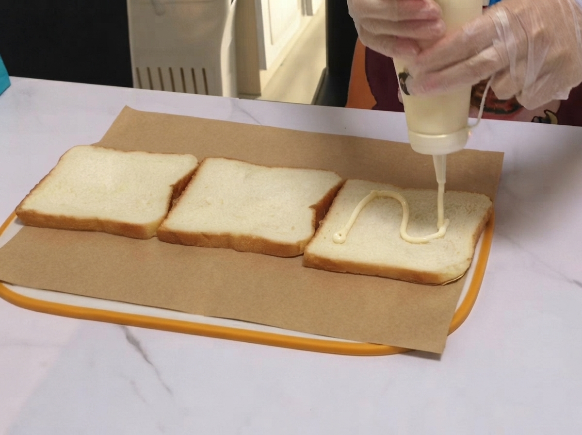 Aderezo blanco siendo aplicado en una línea ondulada a través de una rebanada de pan de molde sobre papel encerado.