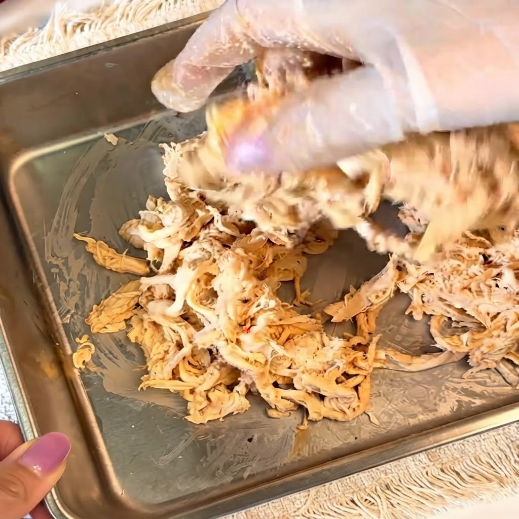 Manos con guantes mezclando pollo desmenuzado sazonado en un recipiente de metal.
