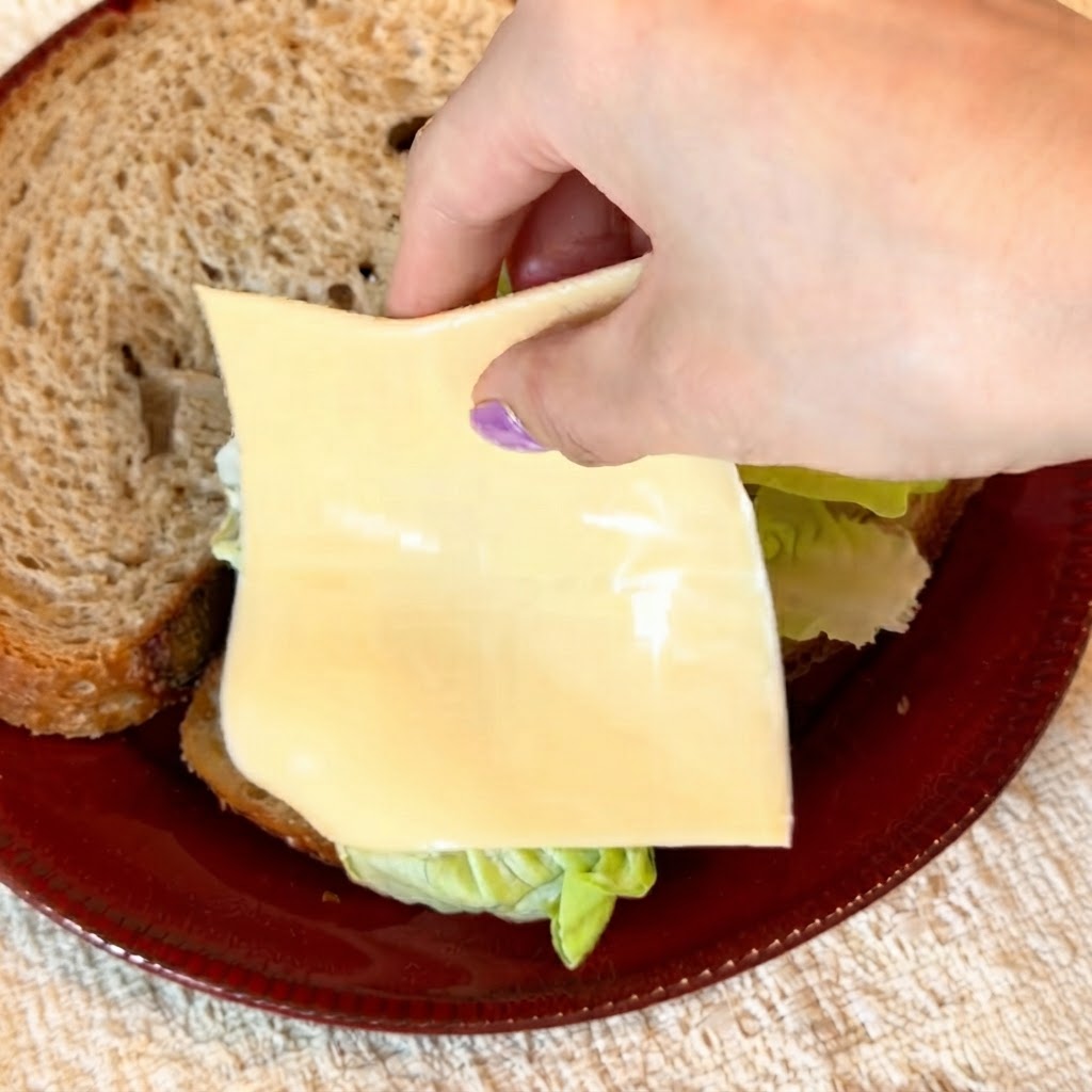 Dos rebanadas de queso amarillo bajo en grasa siendo colocadas cuidadosamente sobre la lechuga verde crujiente.