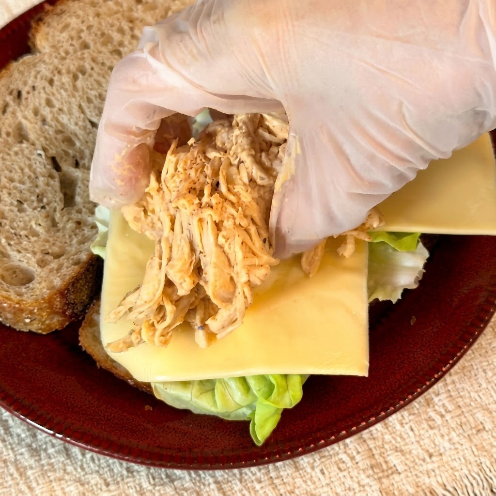 Guantes colocando una porción de pollo desmenuzado sazonado sobre la capa de queso del sándwich.