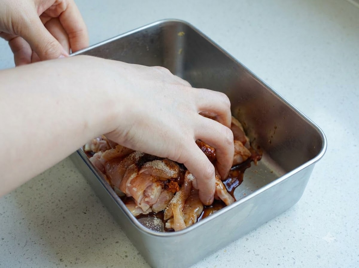 Una mano mezclando trozos de pollo crudo sazonado dentro de un recipiente de metal.