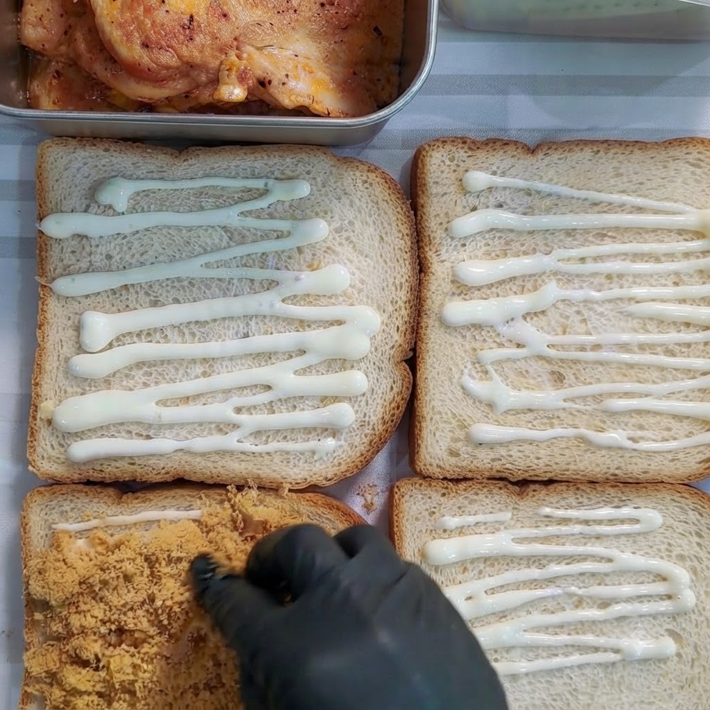Mano con guante esparciendo carne deshebrada de color marrón dorado sobre una rebanada de pan cubierta con aderezo.