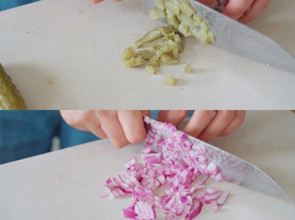 Pepinillos y cebollas rojas siendo picados en trozos pequeños sobre una tabla de cortar blanca con un cuchillo de chef.