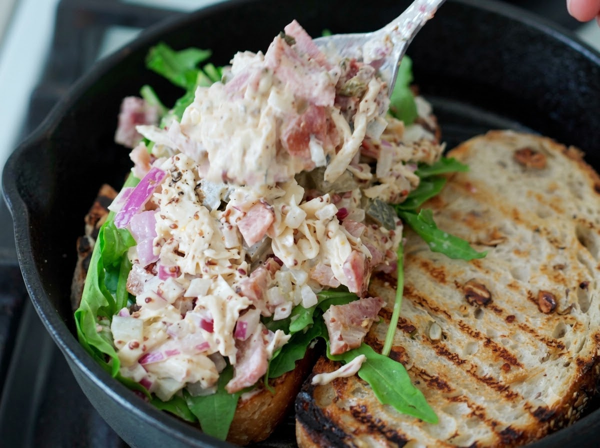 Una cucharada de ensalada de pollo desmenuzado siendo colocada sobre la rúcula verde fresca en una rebanada de sándwich de masa madre.