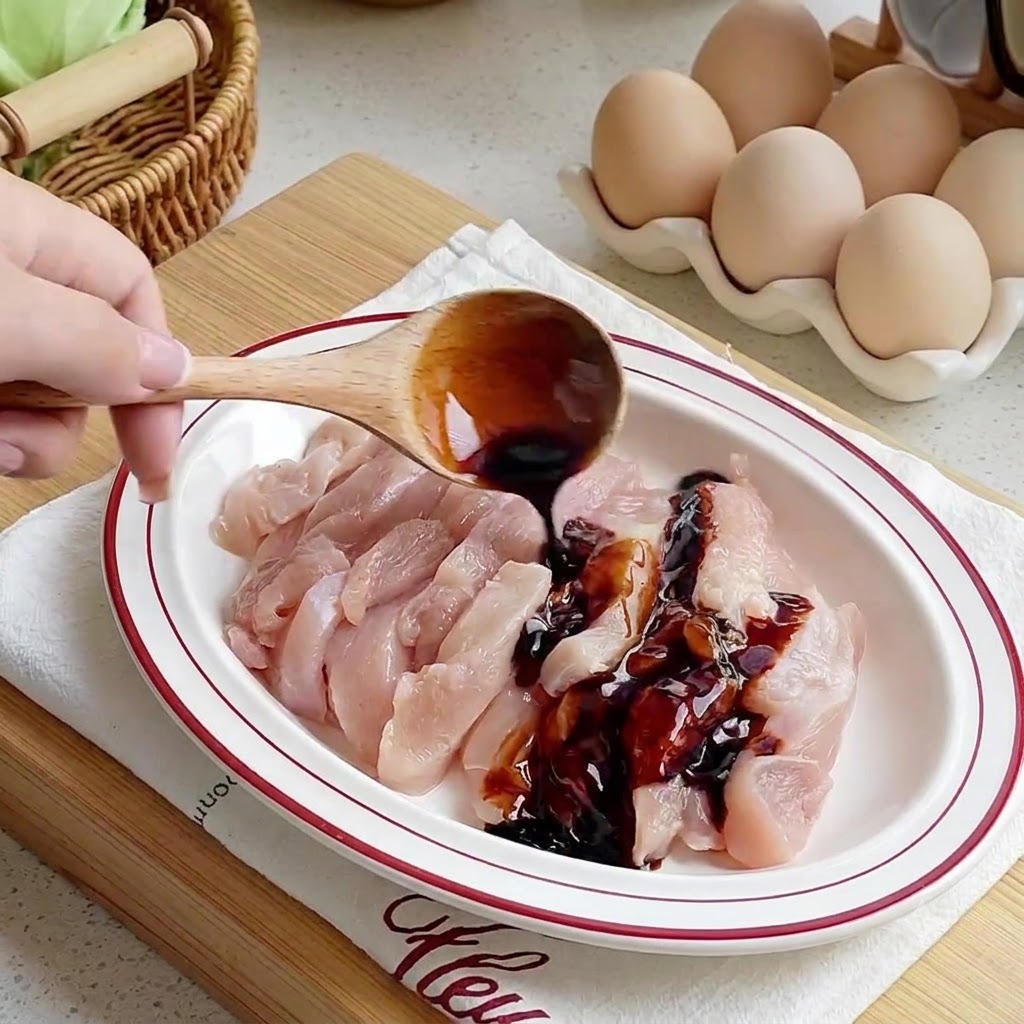 Salsas vertiéndose desde una cuchara de madera sobre tiras de pollo crudas dispuestas en un plato blanco.