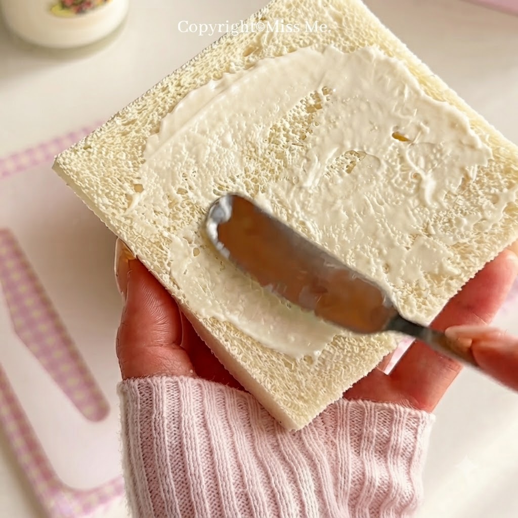 Una mano sosteniendo un cuchillo para esparcir aderezo blanco para ensalada de manera uniforme a través de una rebanada cuadrada de pan blanco.